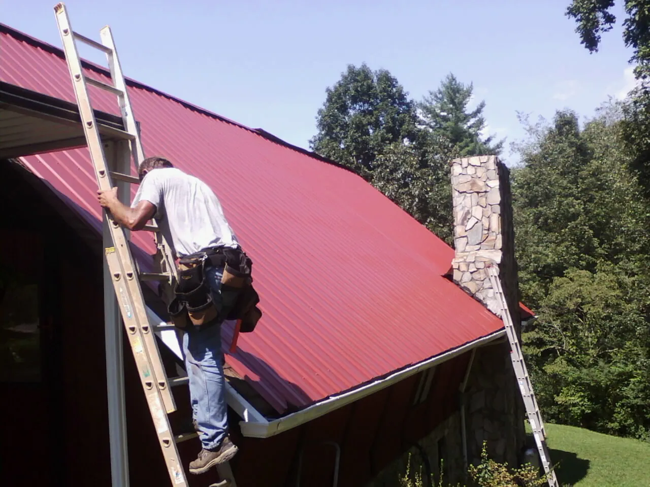 Roofer on a ladder beside a red metal roof and stone chimney — Soffit & Fascia Repair by Roofing Brothers in Flushing